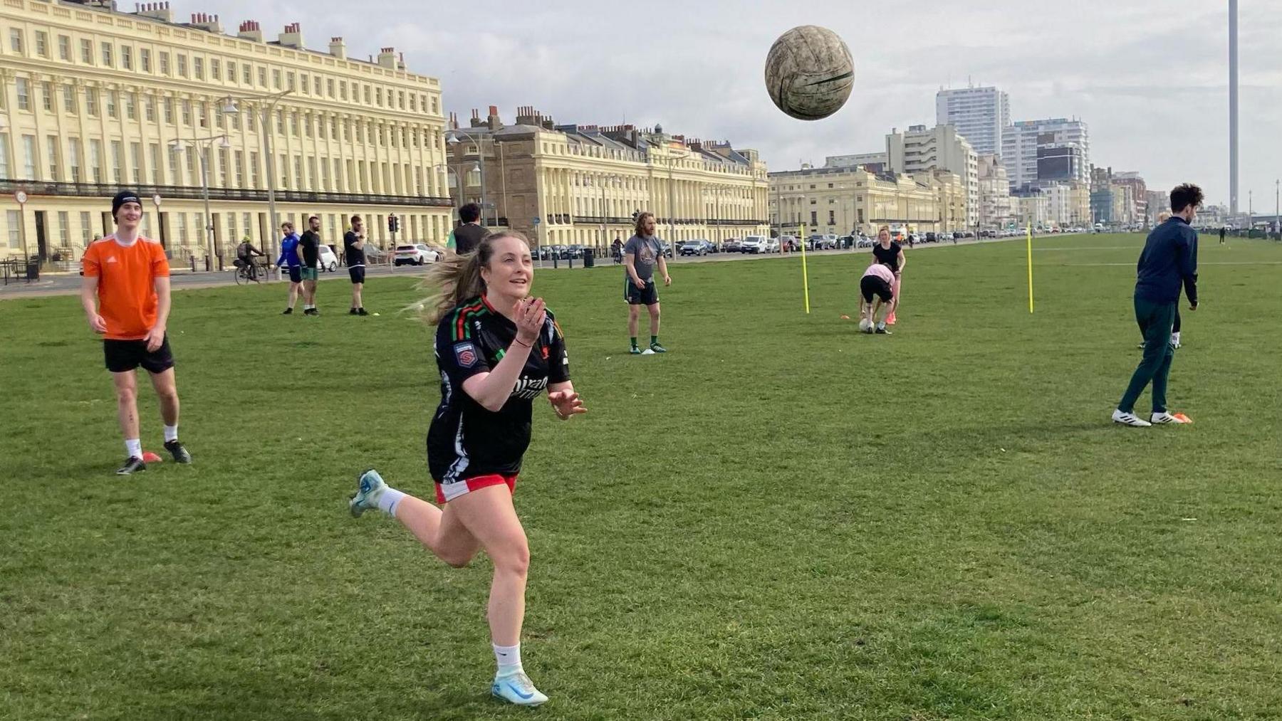 A woman wearing a black t-shirt, red shorts and white football boots runs toward a Gaelic football flying through the air on Hove Lawns. She has her arms reached out to catch it as it is being thrown to her. Other players can be seen stretching, performing drills and setting up a make-shift goal. Hove's iconic cream Regency buildings stretch out across the A259 in the background of the image.