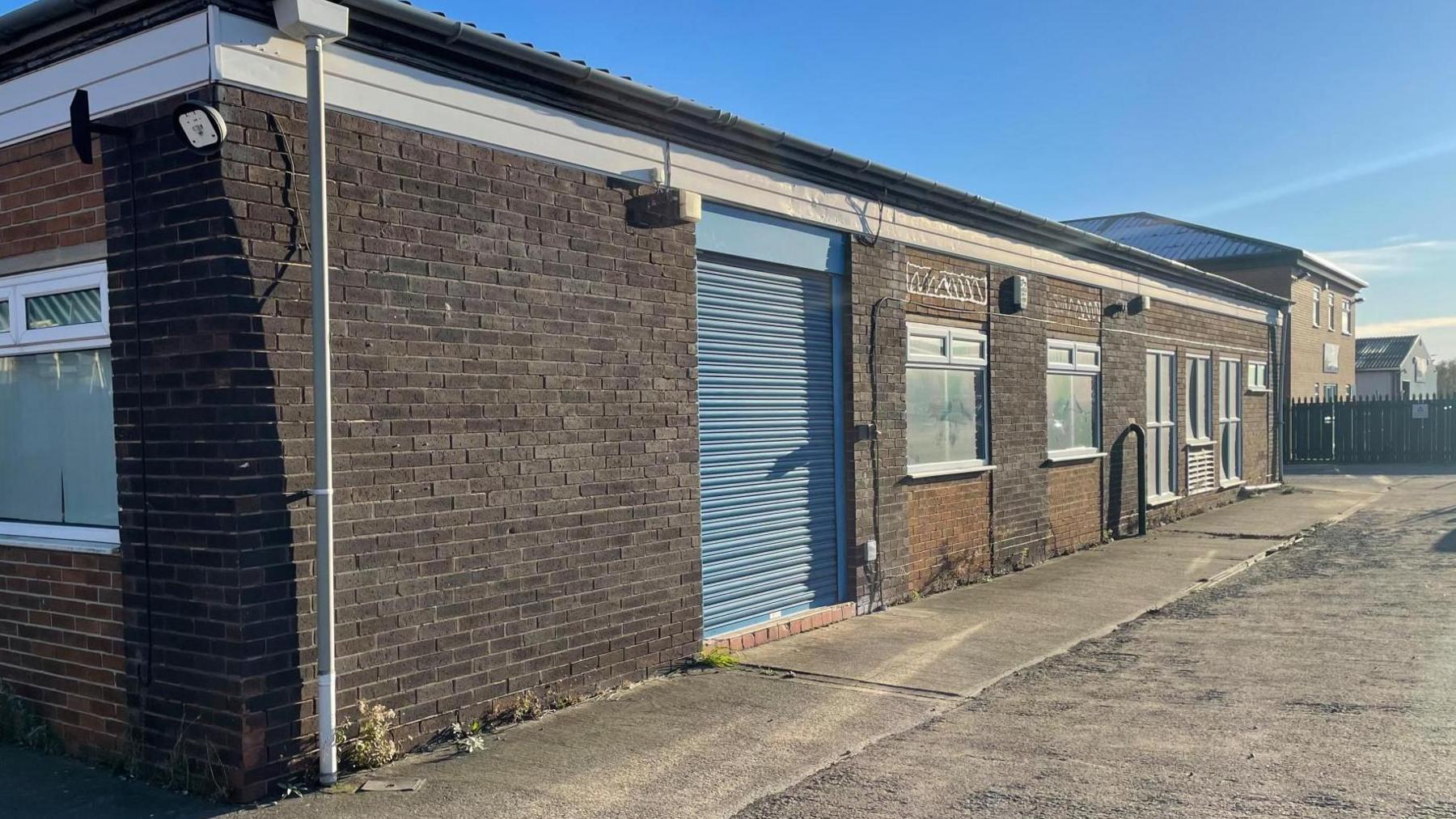 The exterior of a plain brick industrial unit seemingly empty. The blue shutters are down.