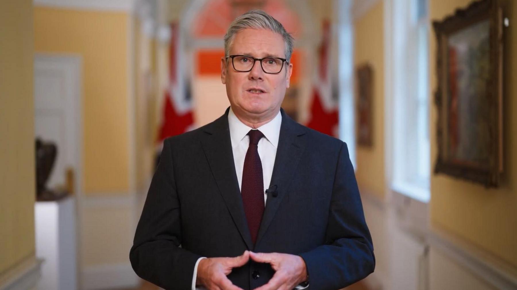 Prime Minister Sir Keir Starmer wears a suit and stands in a yellow corridor with two British flags behind him. The photo is from the video in which he announced the UK would recognise Palestinian statehood.