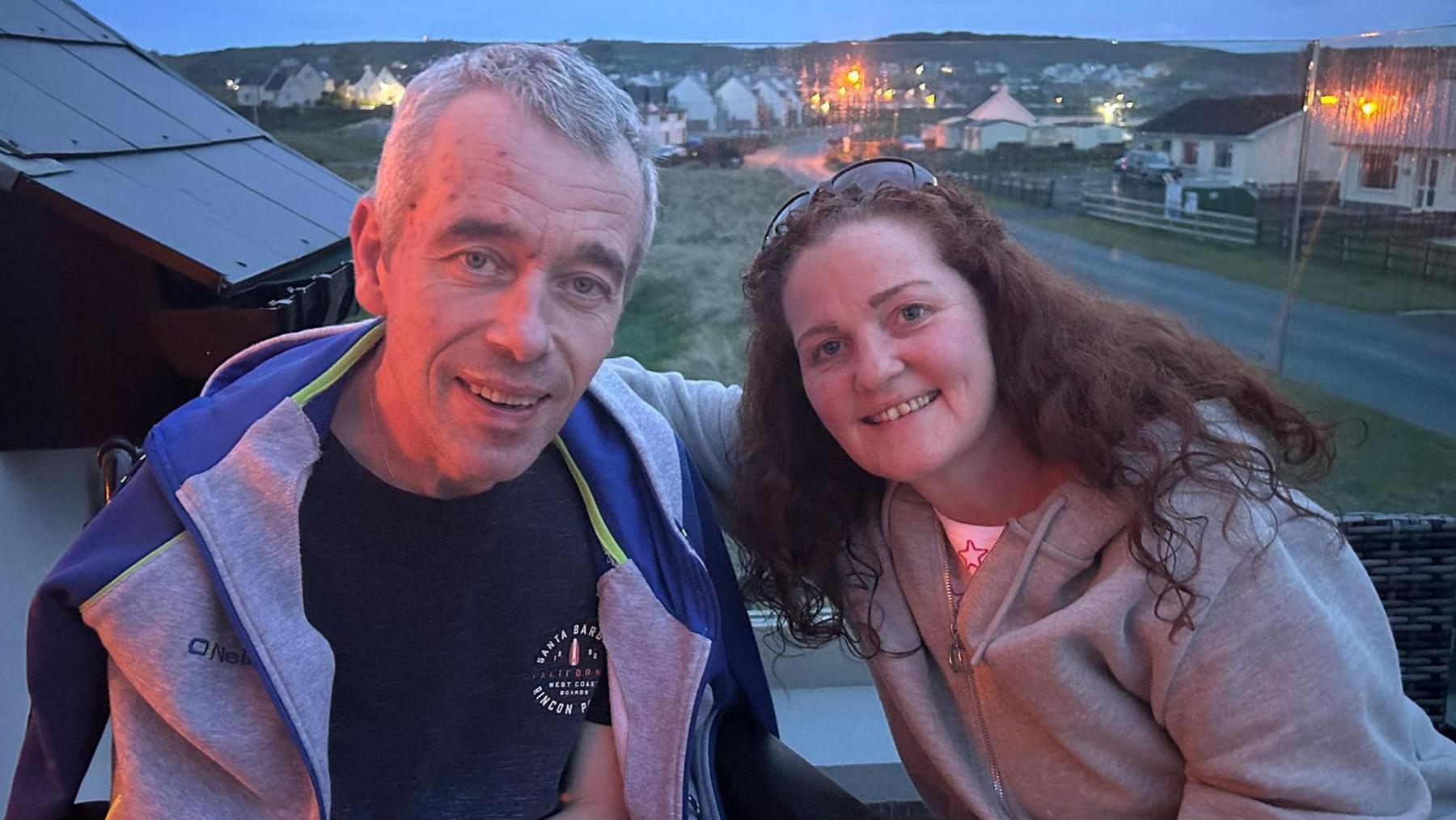 John and his wife Debbie are sat on a balcony overlooking a number of houses and a grassy field. John is wearing a blue and grey hoodie with navy T-shirt and Debbie is wearing a grey hoodie with sunglasses rested on her forehead. 