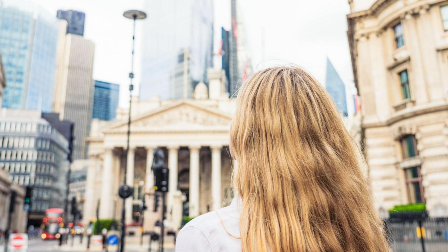 Rear view of a young woman looking up towards the towers in the heart of the City of London, the UK's key financial district, with the Bank of England at left.