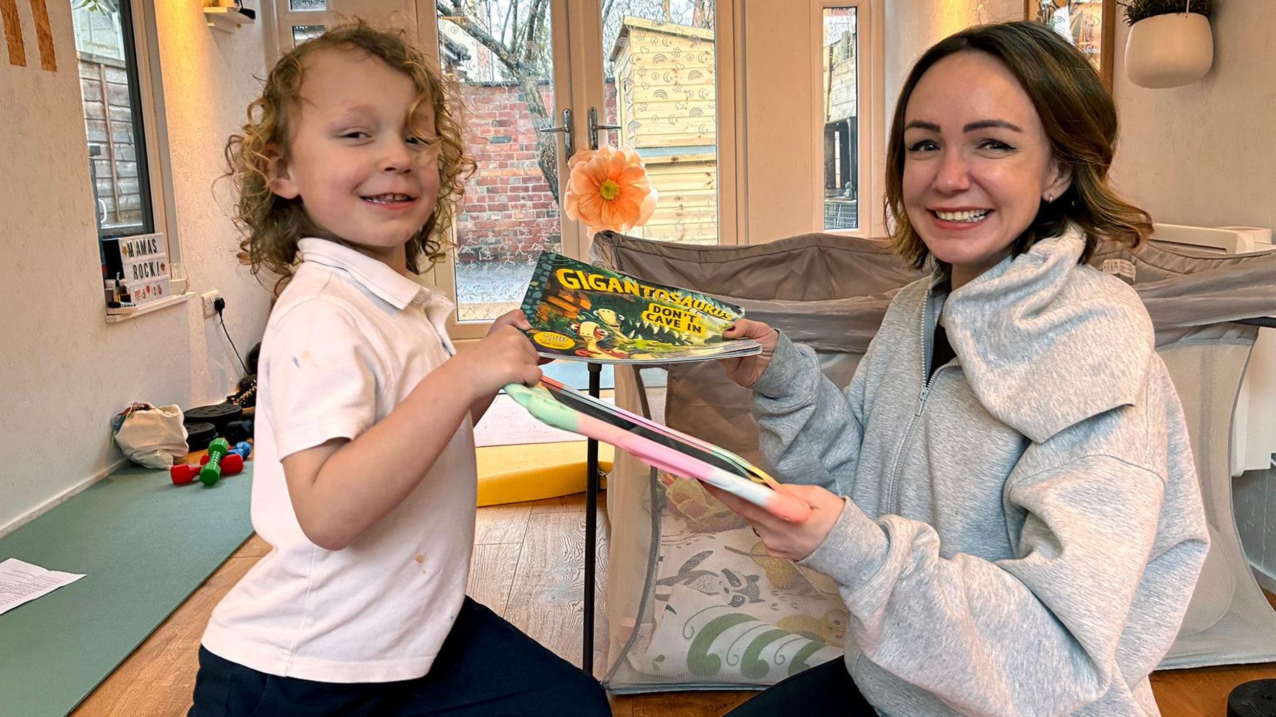 Romi, who has blond curly hair and is wearning a white t-shirt, is passing a book to his mum, Alexis who has short brown hair and is wearning a grey hoodie. She is passing a digital device with a colourful cover back to him and both are smiling.