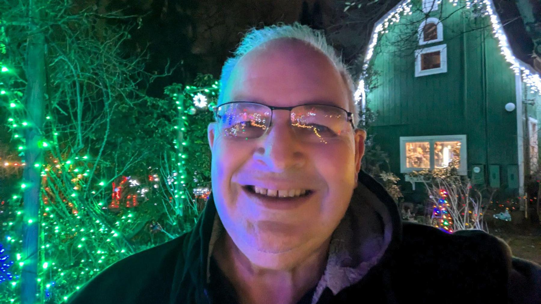 A selfie of an older man with grey hair and clear-framed glasses. He is wearing a black coat and is standing in front of a house that is decorated with Christmas lights. 