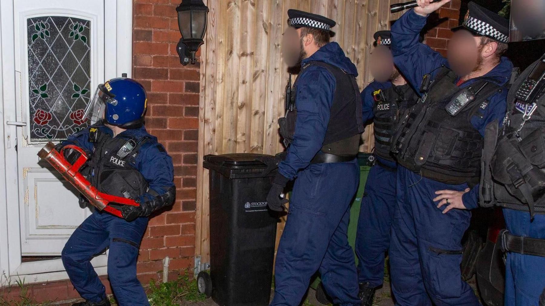 An action shot of a police raid on Moxham's home. Police are dressed in blue uniforms and wearing police hats. One officer is wearing a helmet and is attempting to break down the front door to the property.