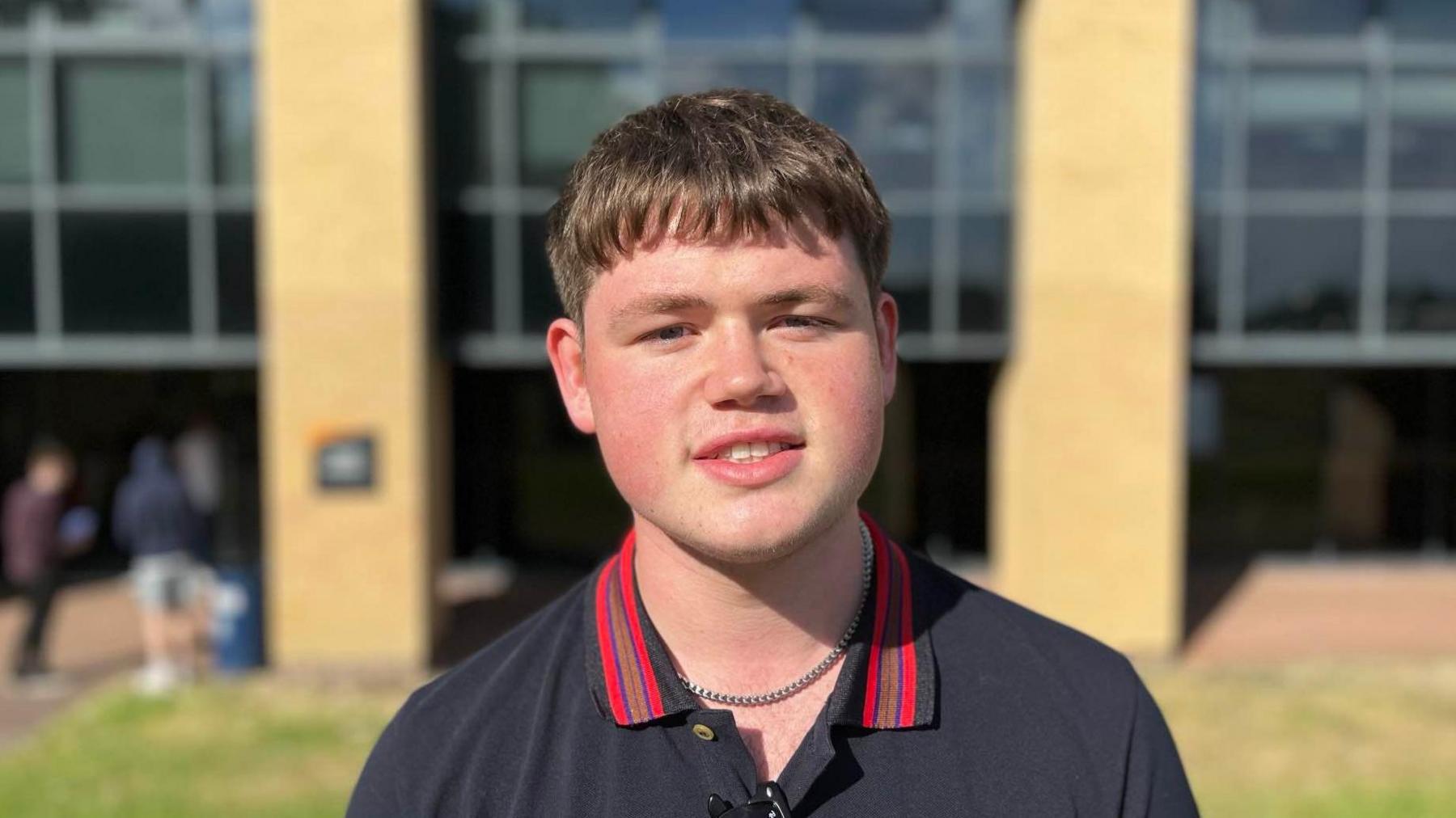 A boy with short brown hair. He is white. He is smiling at the camera. He is wearing a blue polo shirt with red a red collar. He's stood outside in front of a school building