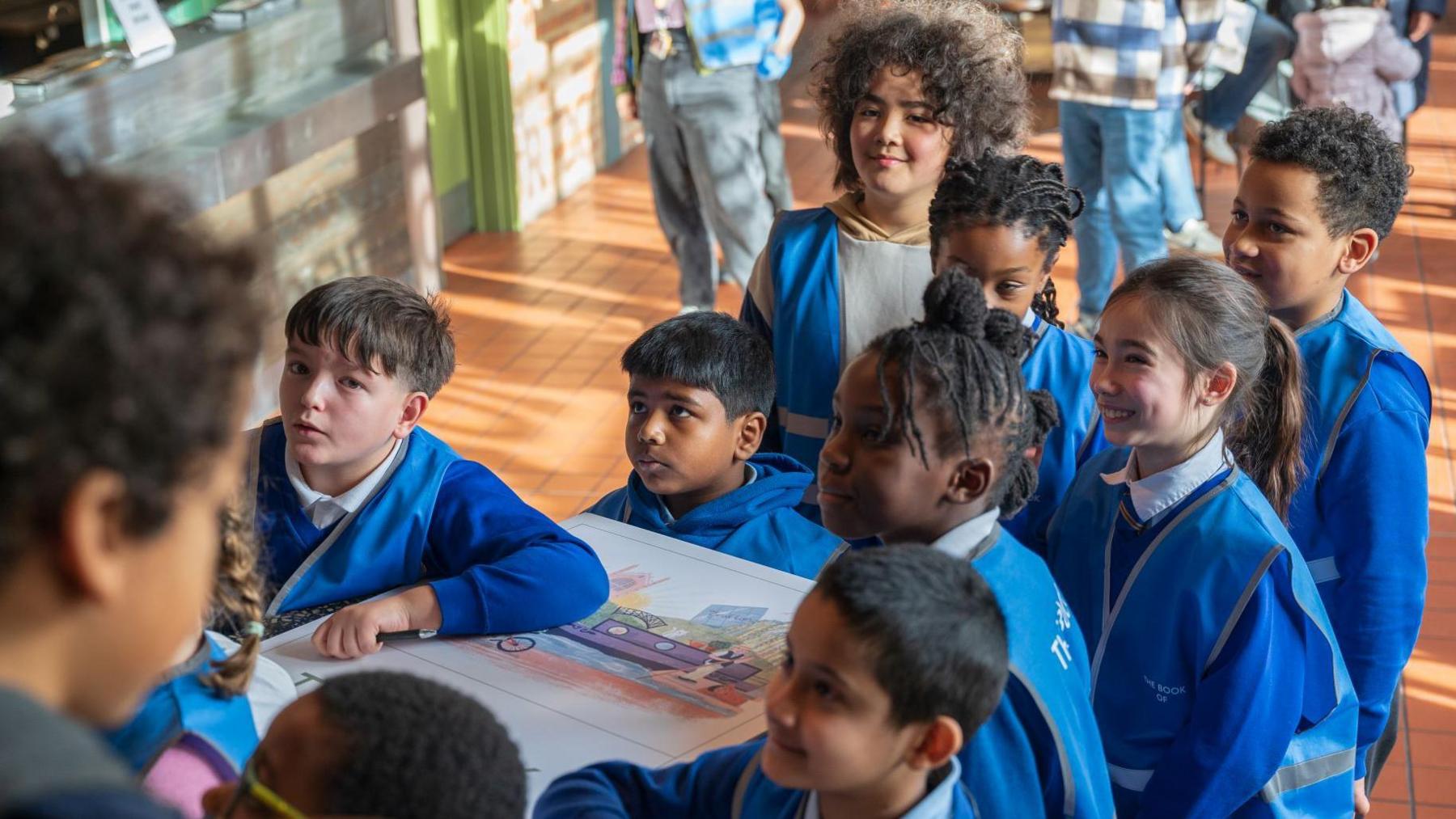 A group of children wearing blue uniform and hi-vis vests all hold a large white version of their book. They are stood in a hall and looking excitedly at something which is out of camera shot.