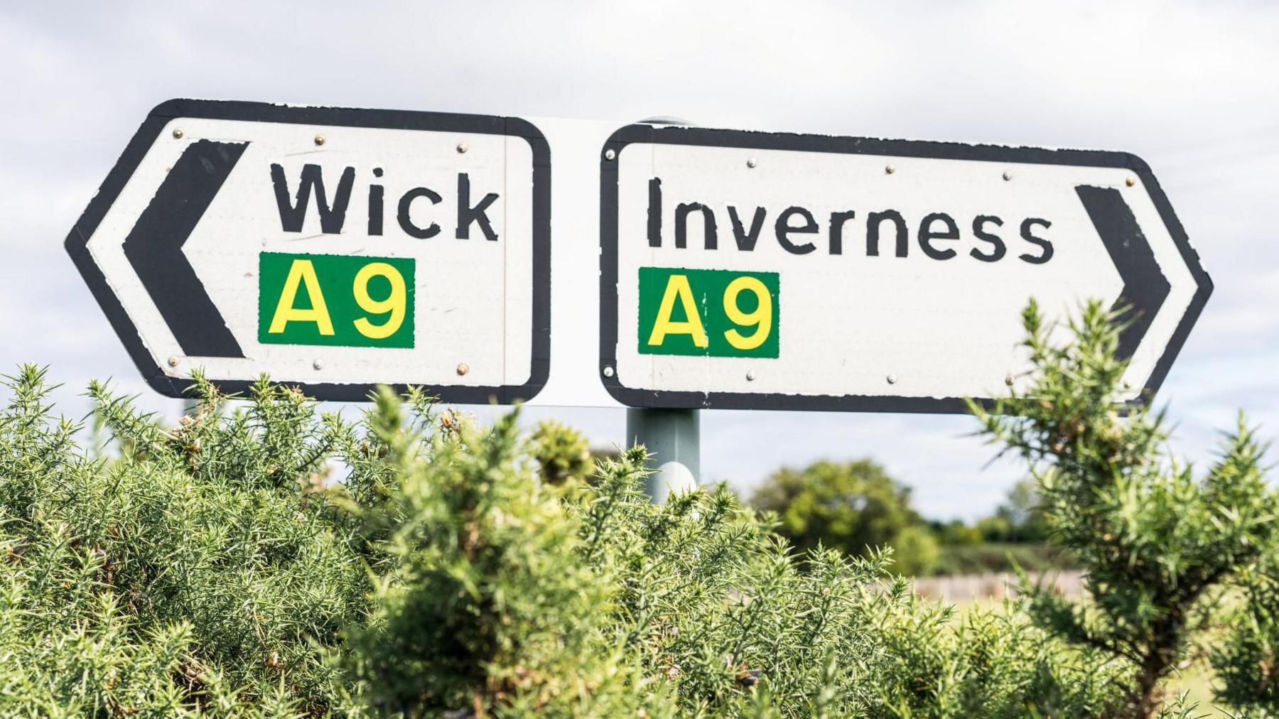Two road signs on the A9 showing the directions to Wick and Inverness. The signs are white with black arrows pointing the way. In yellow in a green box is the word "A9". In black letters are the names "Wick" and "Inverness".