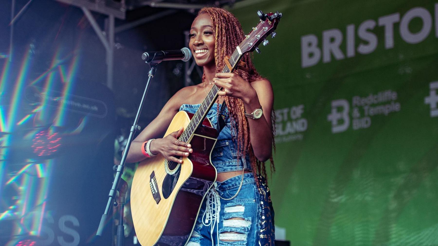 Laura Dia playing guitar on stage at Bristol Harbour festival. She is smiling at the crowd and is wearing a ripped denim outfit