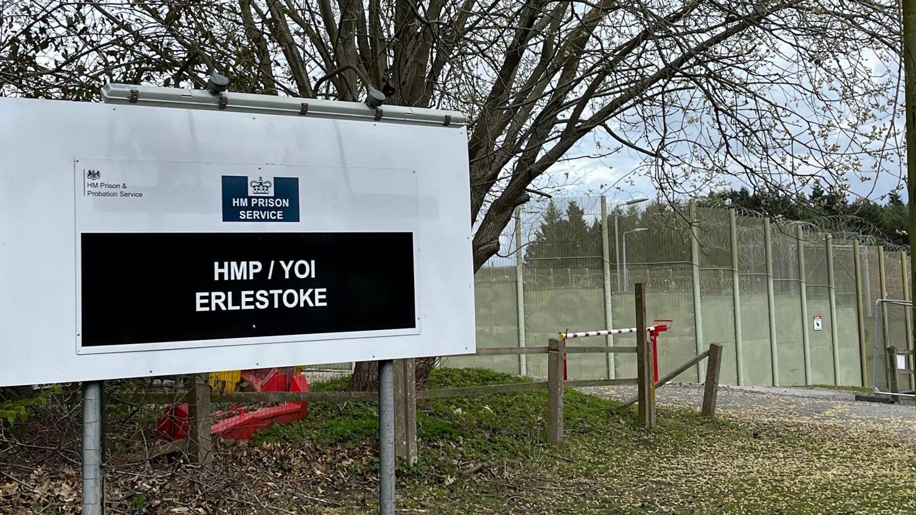The exterior of HMP Erlestoke with a large sign outside and high barbed-wire fences. There is a tree behind the white and black sign, which says HM PRISON SERVICE and HMP / YOI ERLESTOKE.