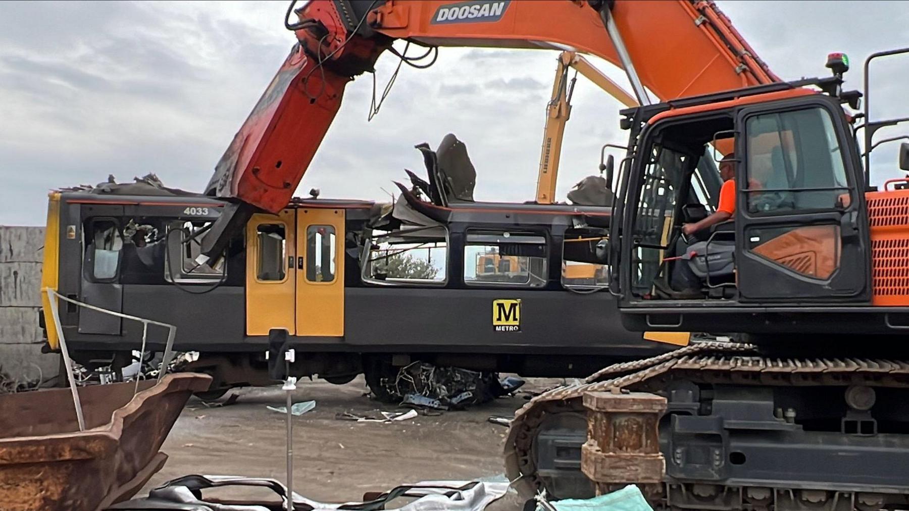 A former Tyne and Wear Metro carriage in its black and yellow livery being dismantled for scrap. A giant hydraulic grabber, operated by a man in a high viz jacket, is crushing the front windows. 