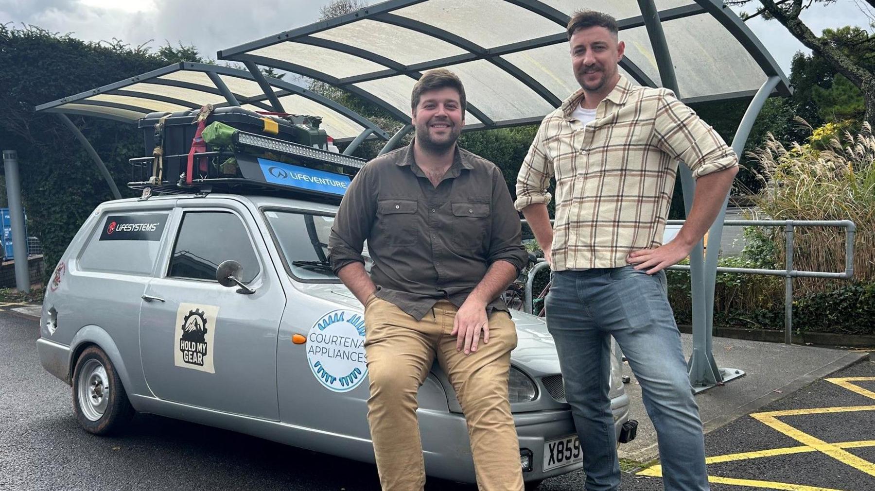 Two men in front of a Reliant Robin vehicle. The man on the left is wearing a chequered shirt and blue jeans, he is holding his hands on his hips. The man on the left is sitting on the bonnet of the vehicle. He is wearing beige chinos and a brown shirt. The car is silver and has a number of stickers on its chassis. There is a roof rack on the top of the car.