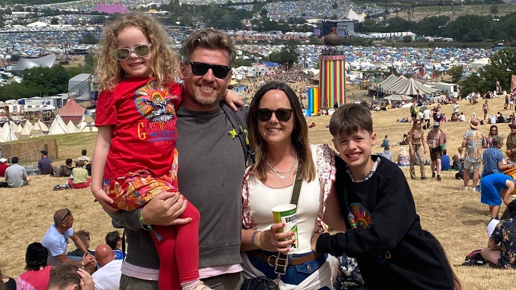 Kelly, her husband and children at Glastonbury Festival. Her husband is second from left and wearing a grey t-shirt and shades, holding their young daughter in his arms, she is wearing a red Glastonbury t shirt and red leggings. Kelly is wearing a white t shirt, sunglasses and holding a cup of drink and their son is wearing a dark top. The whole family is smiling at the photographer with some bright rainbow tents in the background.