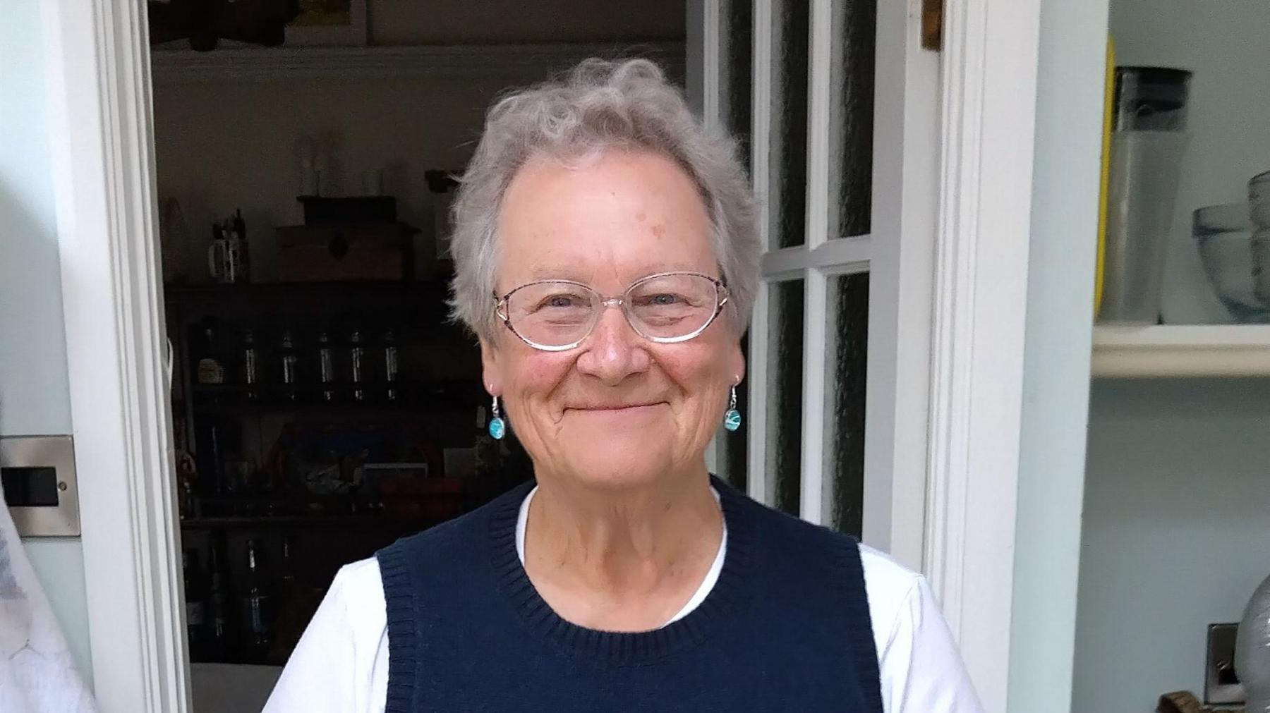 Sally Wainman wearing glasses and a blue and white top. She is standing in a kitchen, only her head and shoulders are visible.