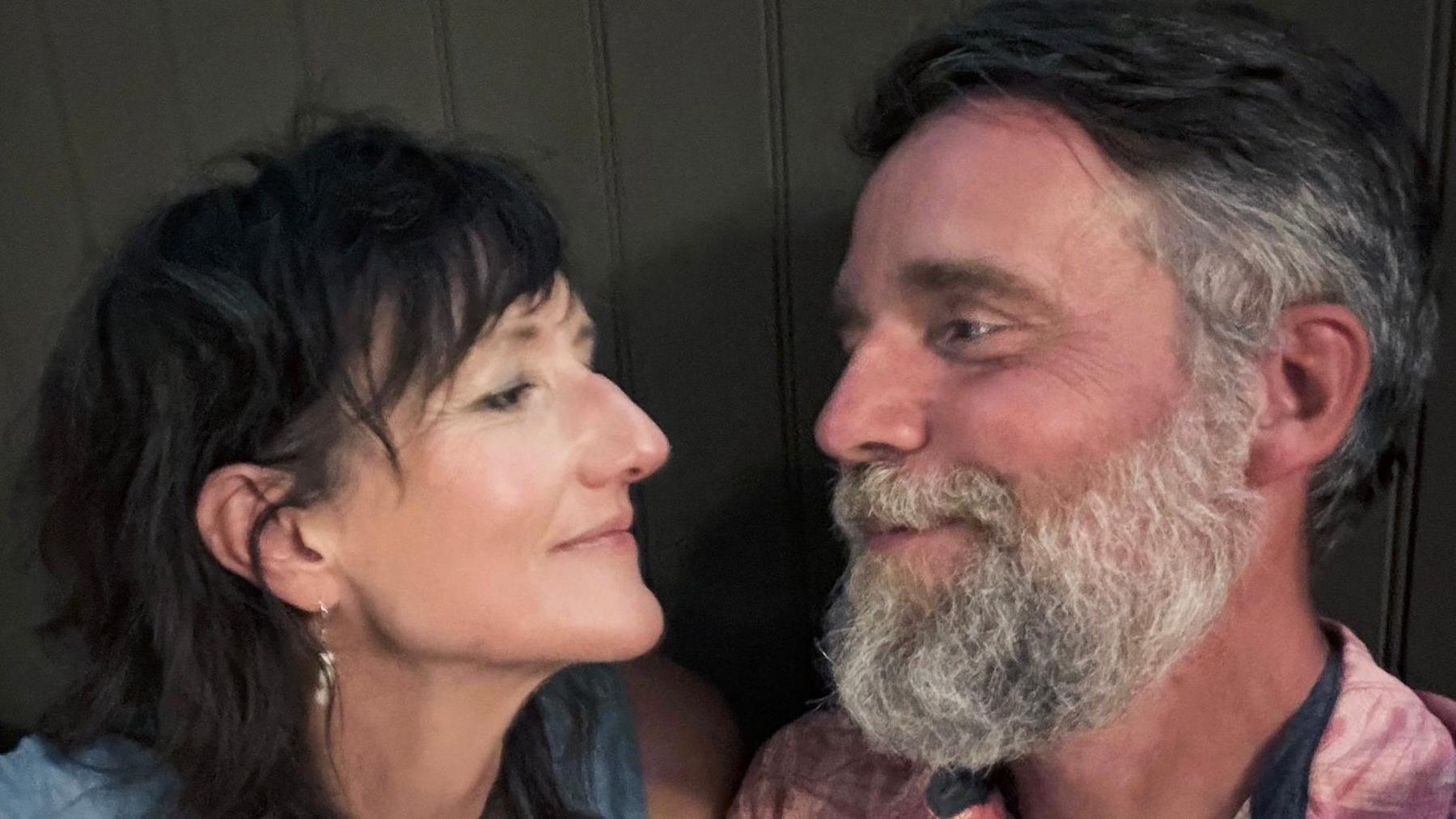 Alice Neal looks at her husband Jeremy. She has short black hair and wears earrings and a blue vest top. Jeremy has dark hair with some grey in it as well as a thick grey beard and wears a pink shirt.