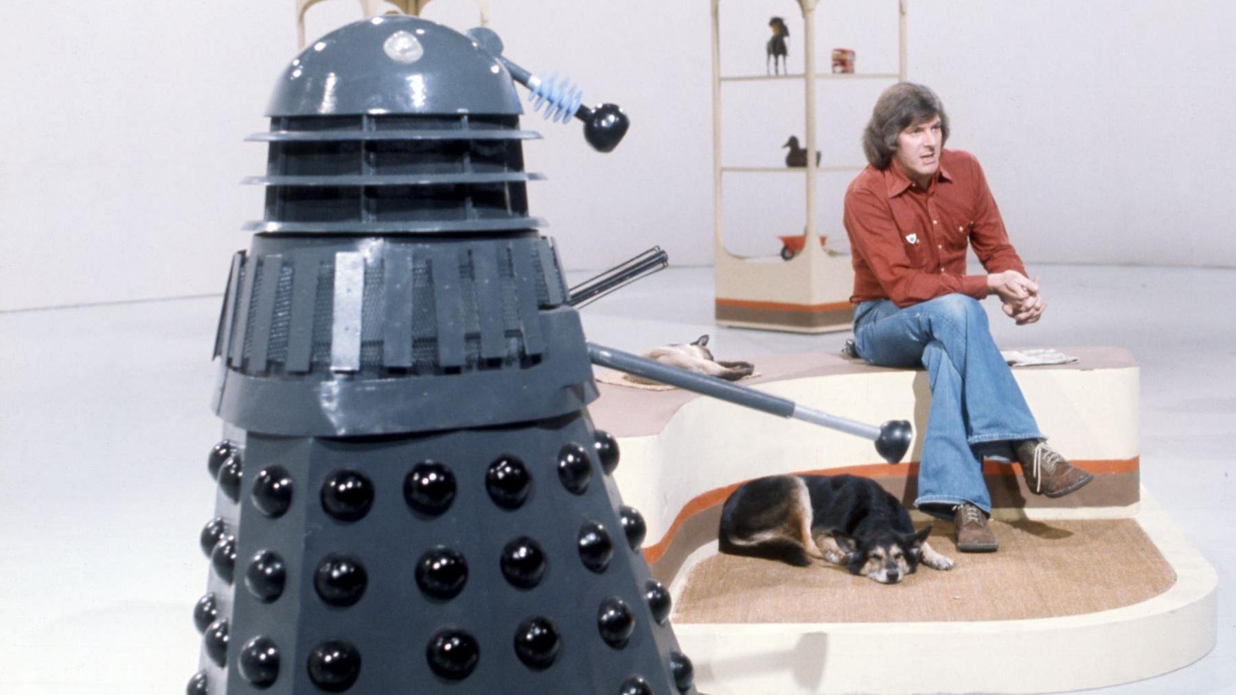 A colour photo of a 1970s television studio, with a primarily white background and floor, with some free-standing shelving in the background. Dominating the photo close up at the left of frame is a Dalek, a conical machine creature, dark grey in colour with black trim. On its right, looking away to a camera out-of-frame, is a man in his late 30s with long, greying dark hair, wearing a red shirt and flared blue jeans. At his feet is a slumbering black-and-brown dog.