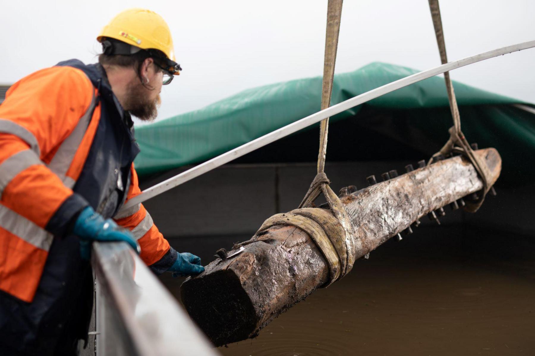 An archaeologist dressed in protective outdoor clothing and a hard hat helps to carefully lower the section of wooden ship into a large water tank.