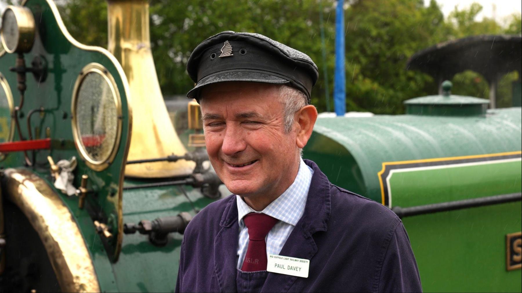 Paul Davey stands on a platform next to a locomotive. He is smiling away from the camera. He wears a train conductor uniform with a hat and name badge. He has grey hair.