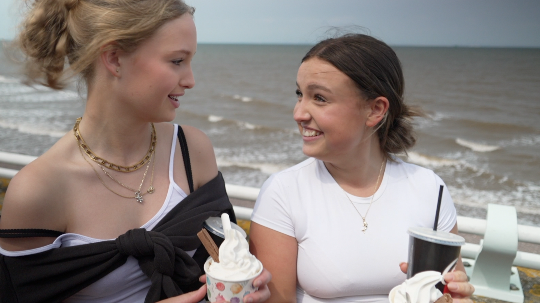 Two teenage girls turn to smile at one another. They are both holding Mr Whippy-style ice creams with chocolate flakes, in tubs. The sea is not far behind them.