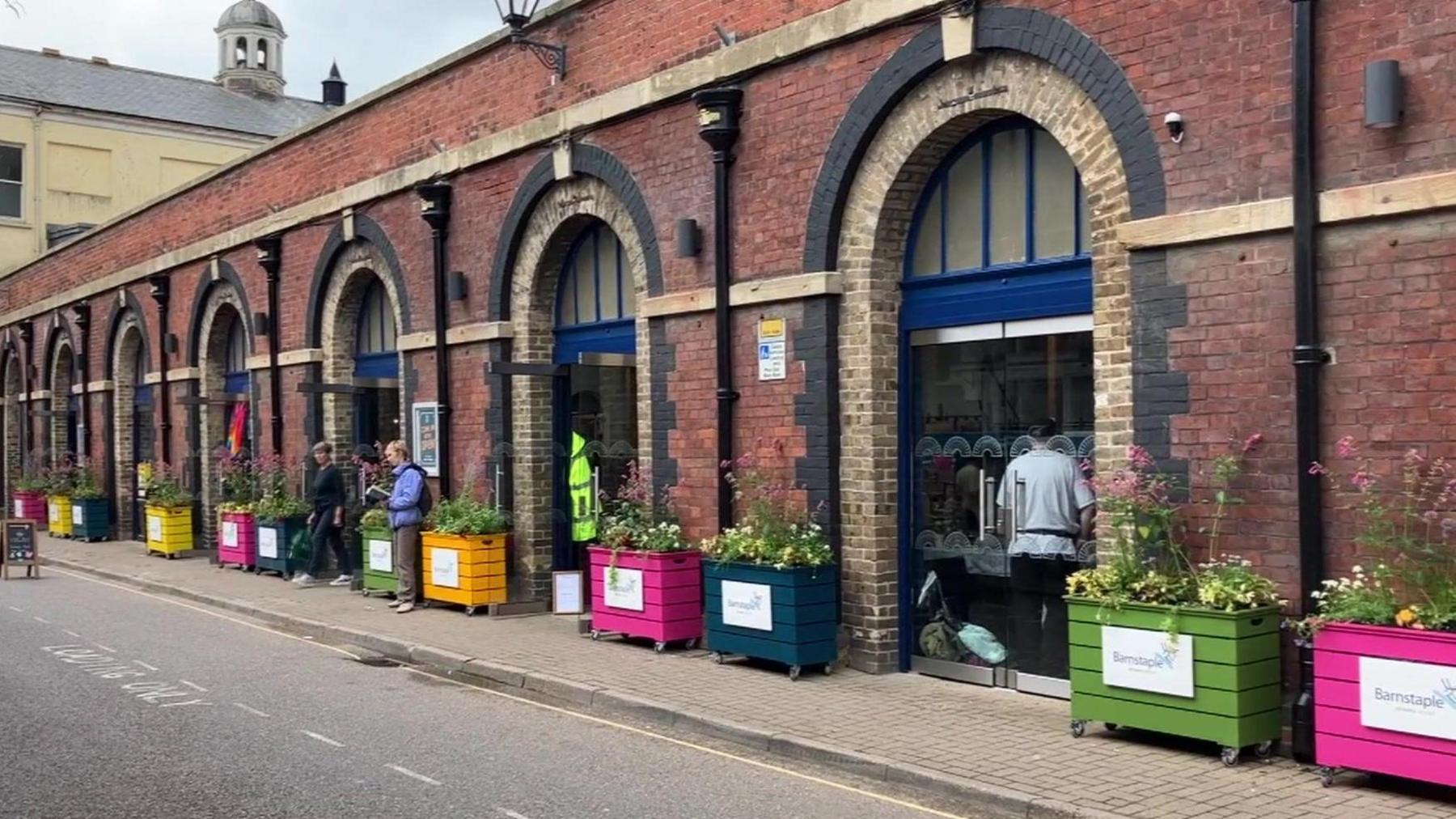 Pannier Market in north Devon - a long one story brown brick building with different coloured potted plant holders, seven entries, grey skies