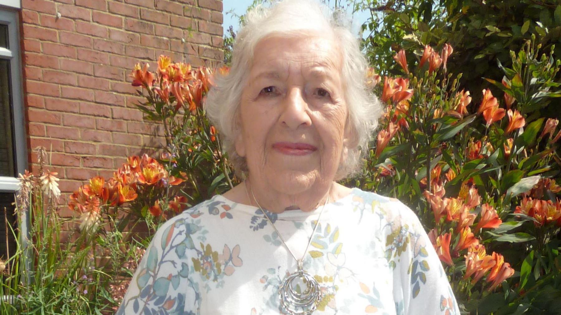 Phyllis Babb. She has short white hair and is pictured outside, in front of some orange flowers. She is wearing a white top with a floral pattern and a silver necklace. She is looking directly at the camera and smiling.