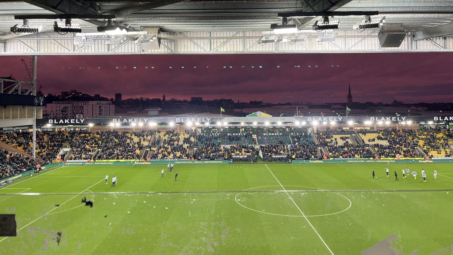 Flood lights lighting up Carrow Road. In the background a purple sky highlights the city skyline