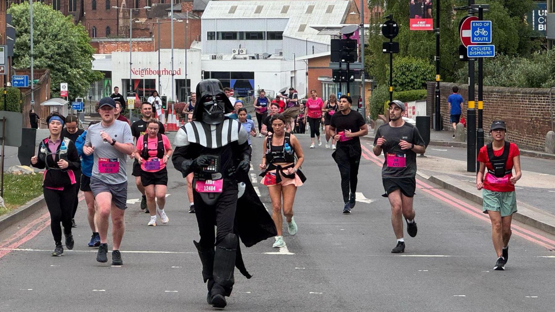 A person dressed as Darth Vader from Star Wars is running in front of other men and women, who are also competing but wearing shorts and T-shirts.