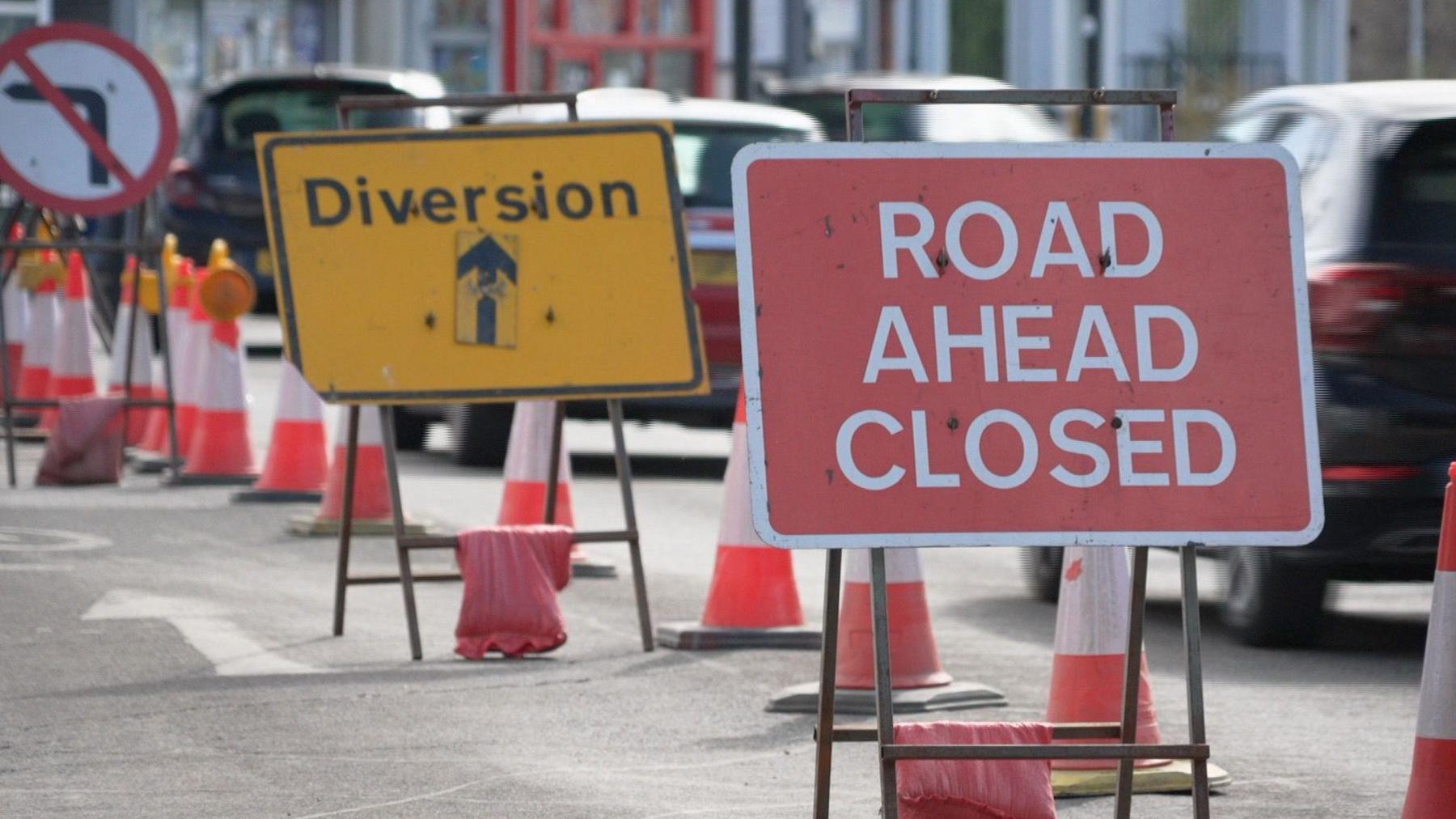 Road closed sign and yellow diversion sign as cars drive past.