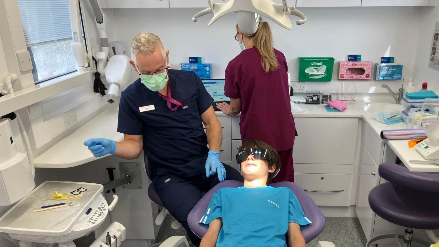 A 12-year-old boy sits in a dentist's chair. He wears protective goggles. The dentist wears scrubs and a mask and rubber gloves. In the background, an assistant prepares the equipment.
