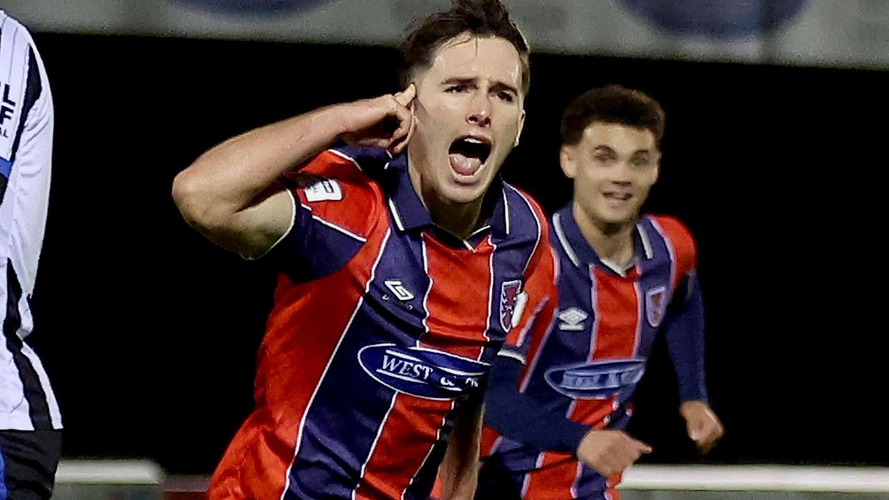 Joe Haigh in red and blue striped Dagenham shirt runs away celebrating a goal by pointing to his head as a team-mate tries to catch up with him