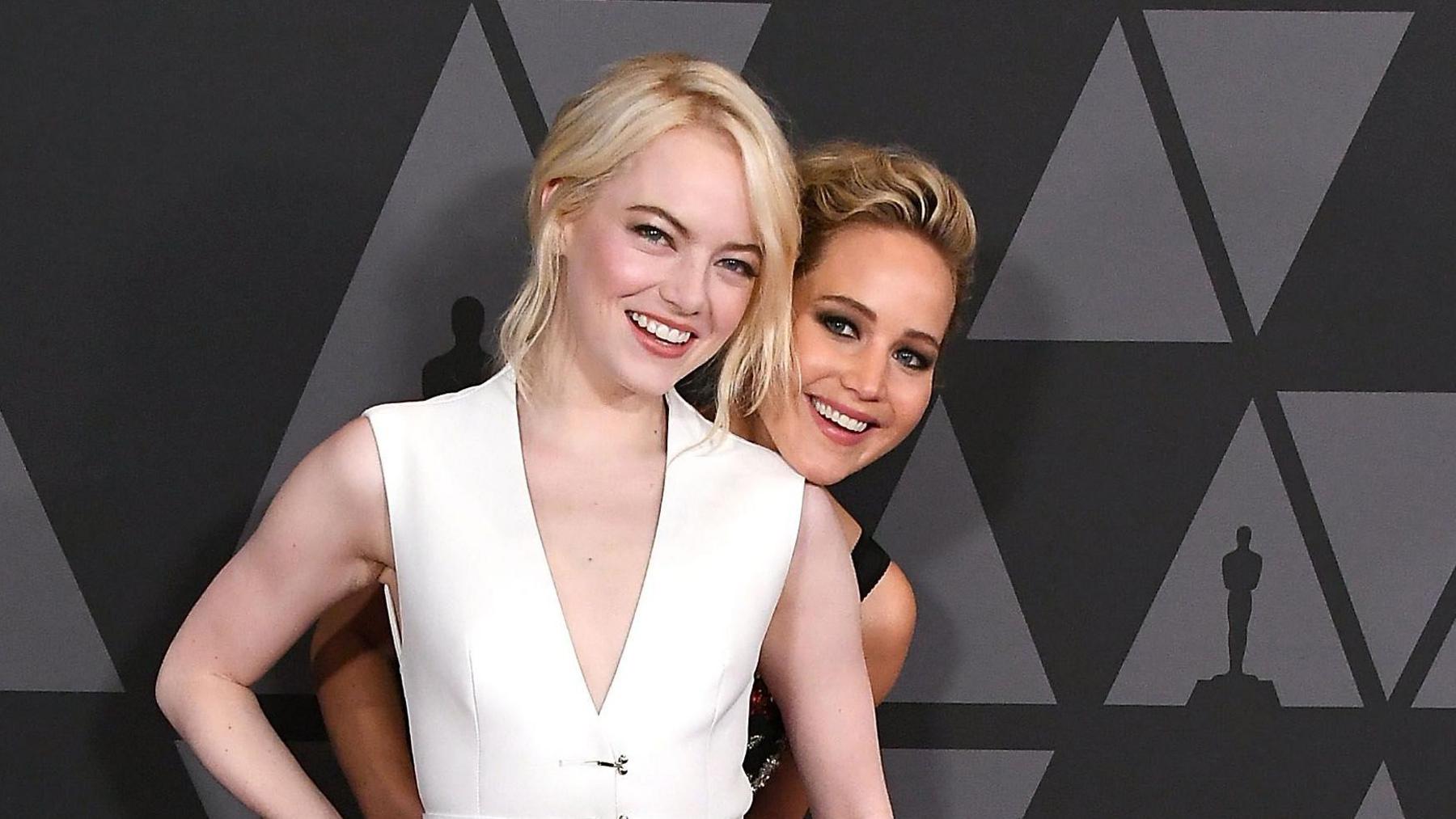 Emma Stone wears a white dress and has blonde hair. Jennifer Lawrence stands behind her.