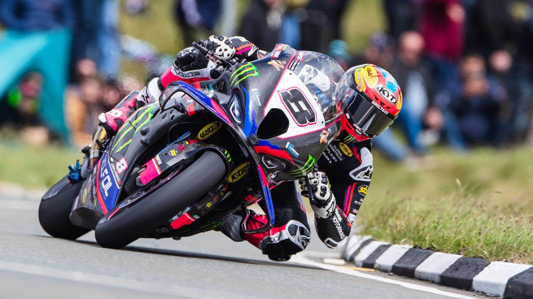 Davey Todd leaning over heavily on his Superbike, which has colourful livery including blue, pink and black, and has the letter eight on the front in black lettering on a white plate. His helmet is blue, red and yellow.