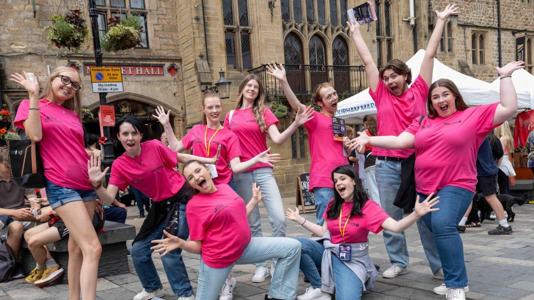 A group of nine people are outside Market Hall in Durham with their arms in the air and joyous looks on their faces. They are all wearing pink t-shirts, which say "Durham Fringe Festival" on the front.