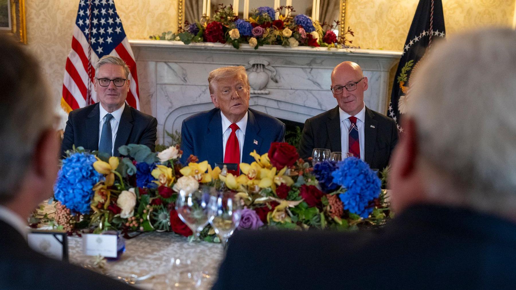 Present Trump attending a dinner with Keir Starmer (left) and John Swinney (right). All three men are facing the camera and are all wearing suits with white shirts and ties. There are colourful flowers and glasses on the table and flowers on a large fireplace behind them. There are also American flags behind them.