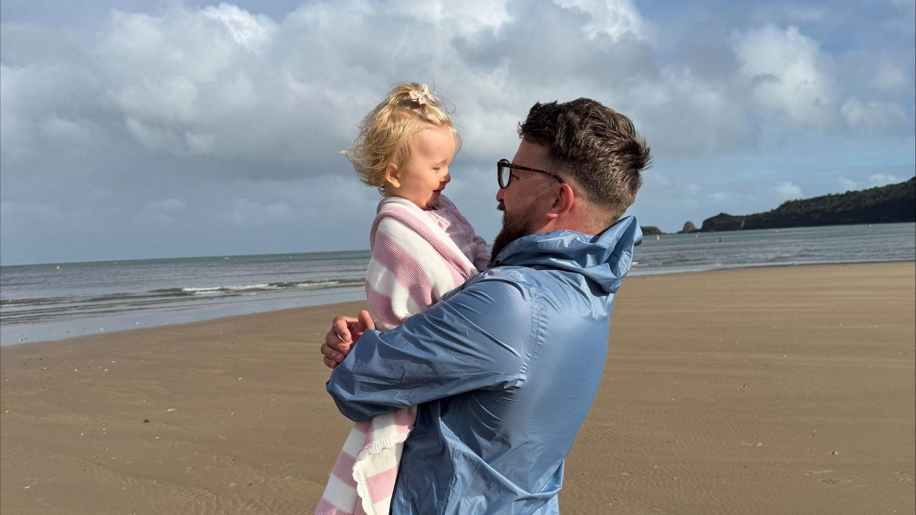 A man holds his daughter on a sandy beach. They are staring into each other's eyes. She is laughing. 