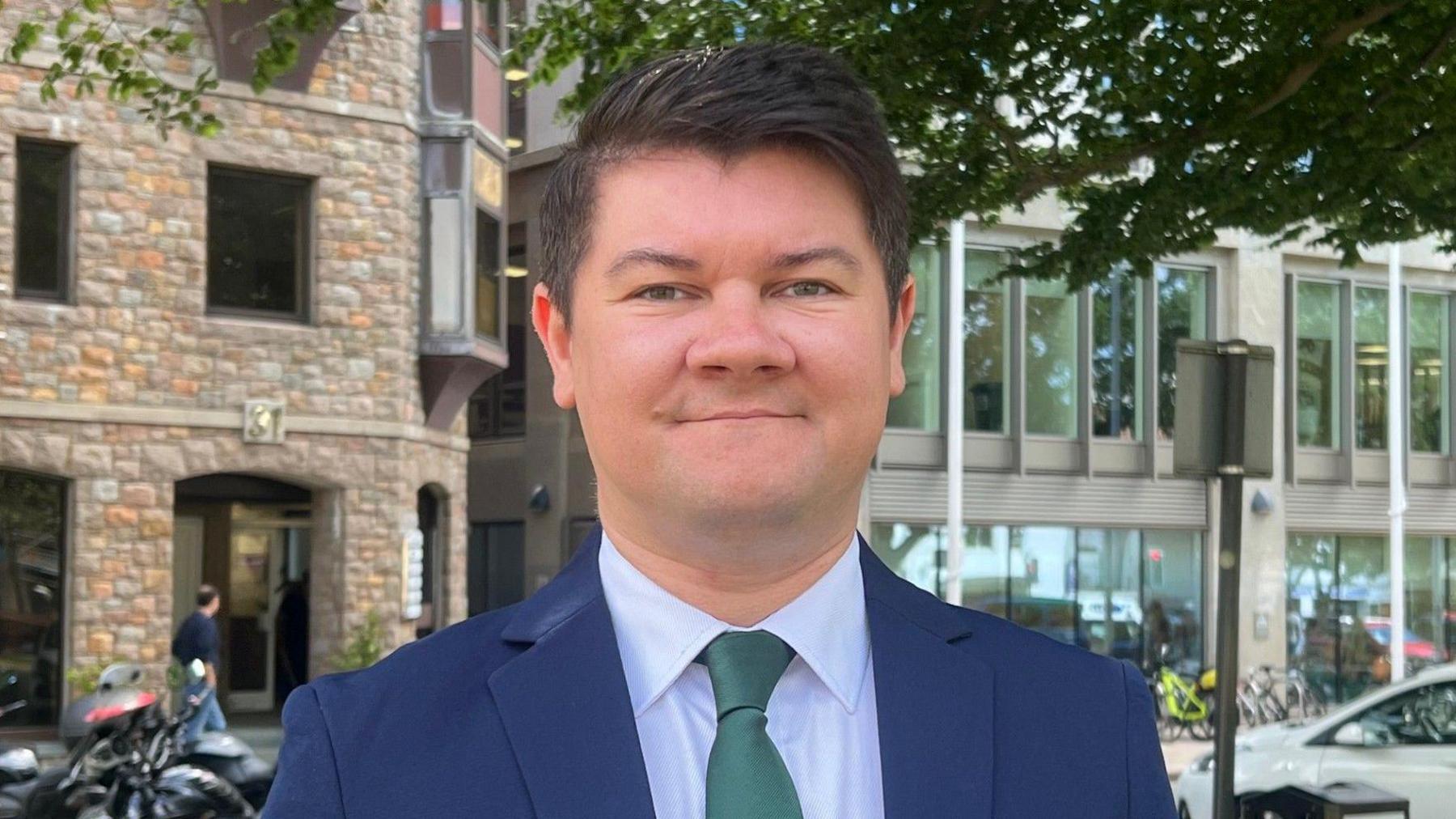 Deputy Sam Mezec is wearing a navy blue suit, a light blue shirt and a green tie