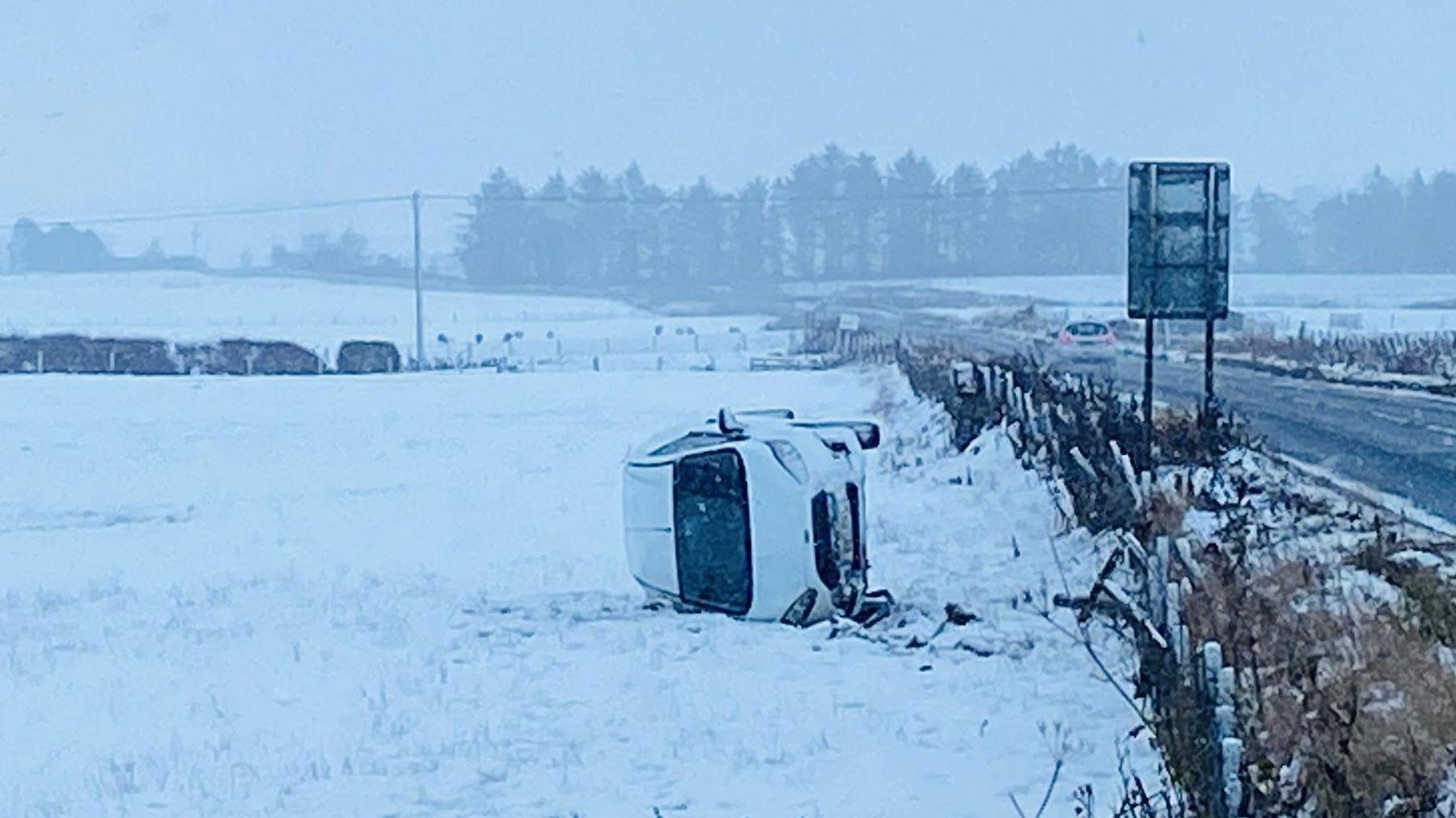 White car on its side in a snow-covered field next to a country road.