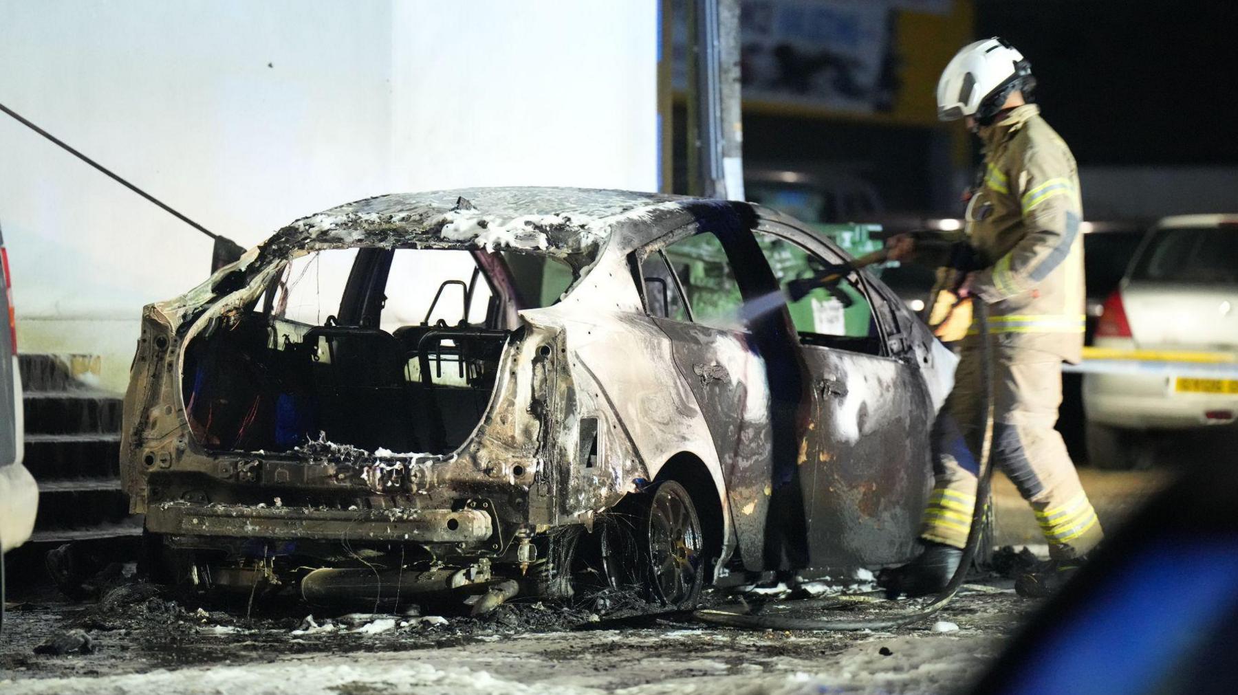 A firefighter spraying a hose into the right rear window of a burned car at night. The photo is taken from behind and to the right of the vehicle