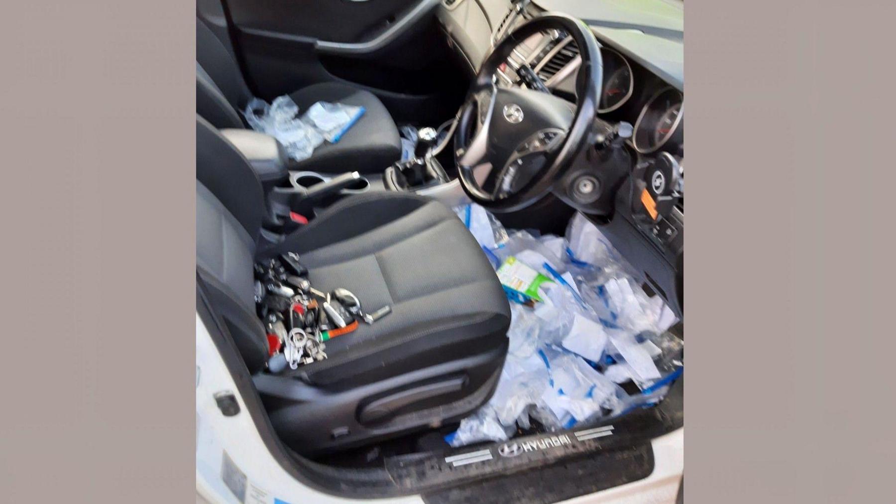 A car is seen with the passenger door open and on the seat are various keys to other vehicles while dozens of plastic bags are visible on the floor