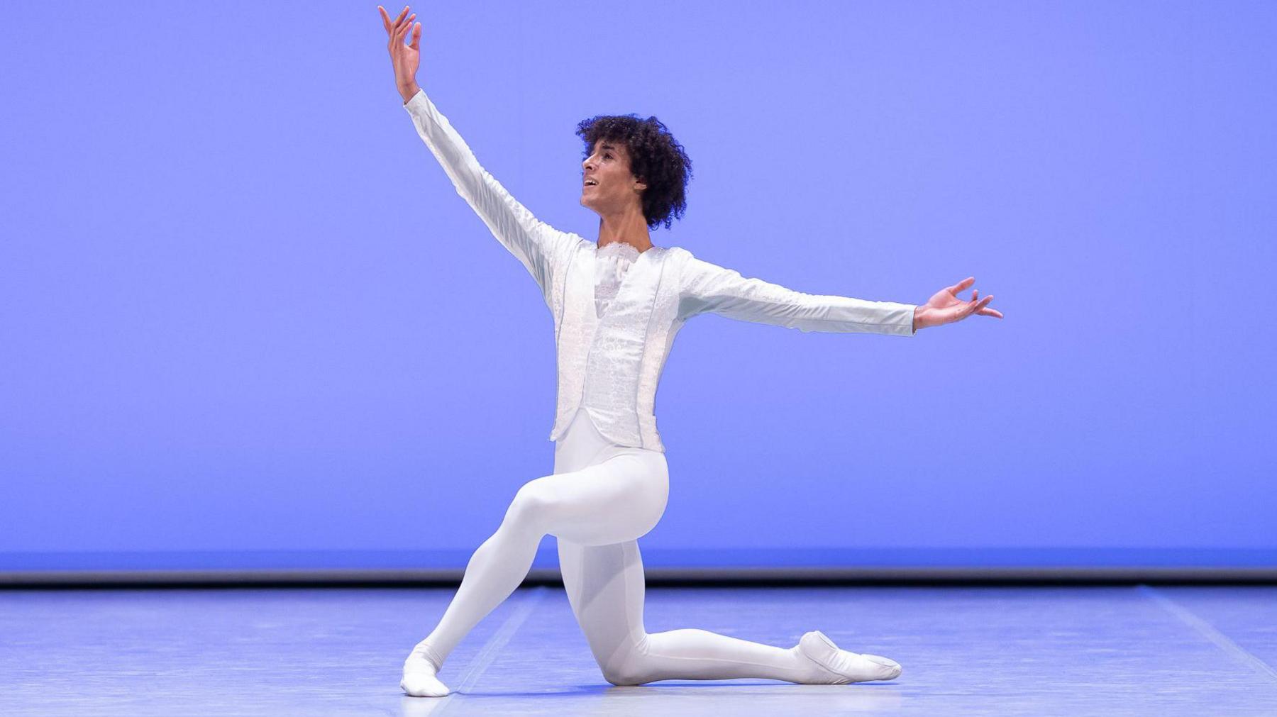 Jakob in a fully white dancers outfit with arms outstretched. He is on a stage, with a blue backdrop. He is smiling. He has short curly dark hair. 