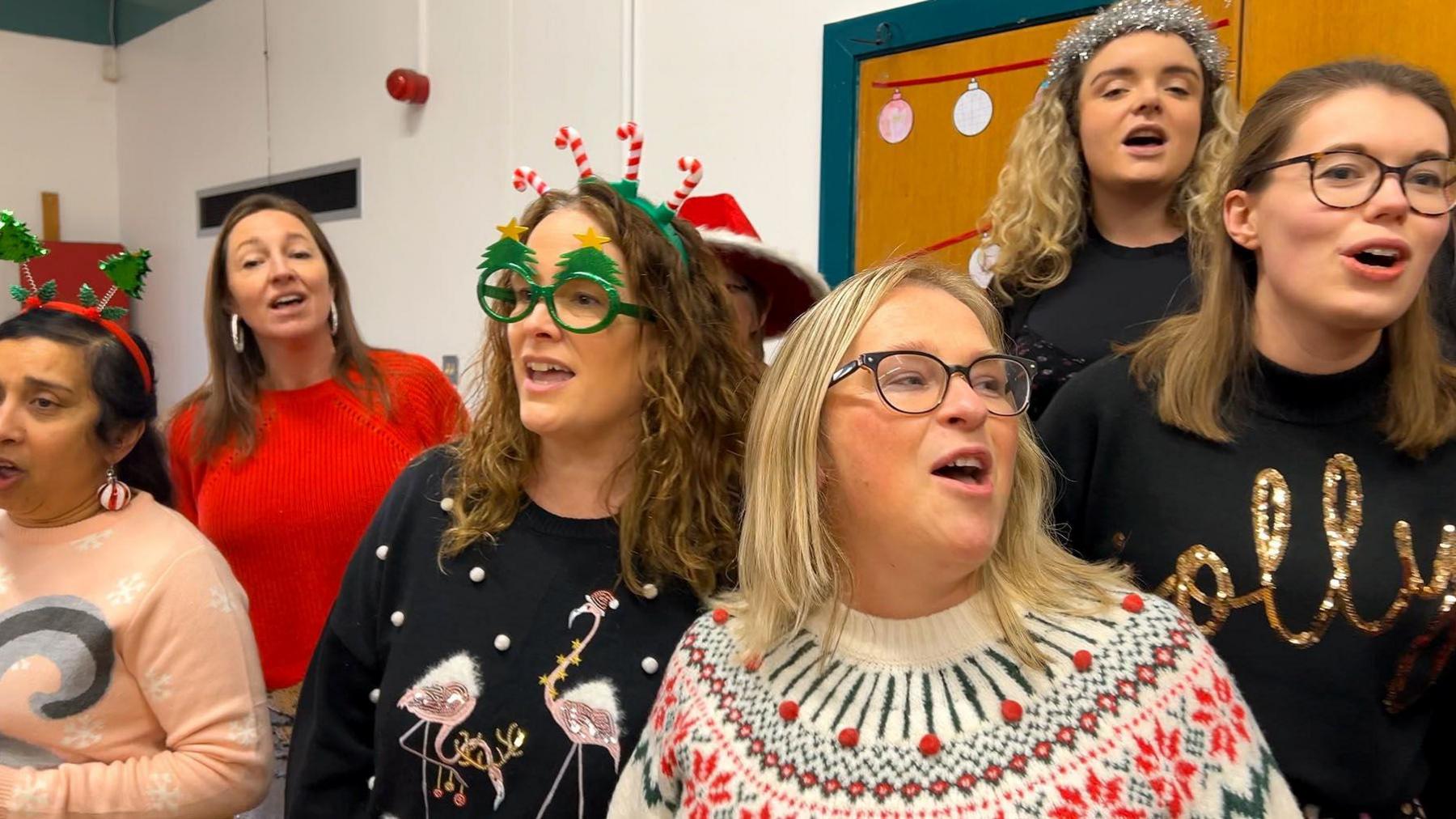 Six women, who are all wearing Christmas jumpers and accessories, looking away from the camera and singing.