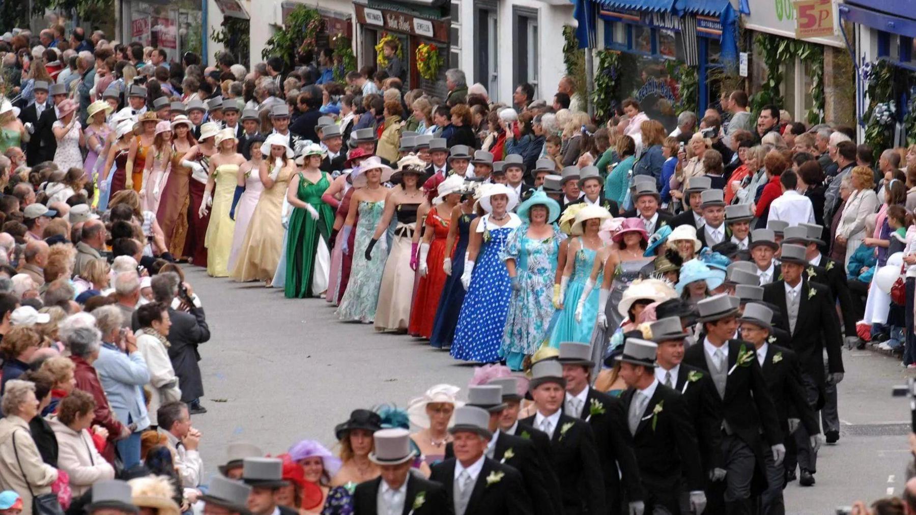 About 30 couples are walking along a street which is lined with spectators. The ladies are wearing brightly-coloured full-length gowns with hats and gloves. The gentlemen are wearing top hats and tails. The shops lining the street are decorated with greenery and flowers.