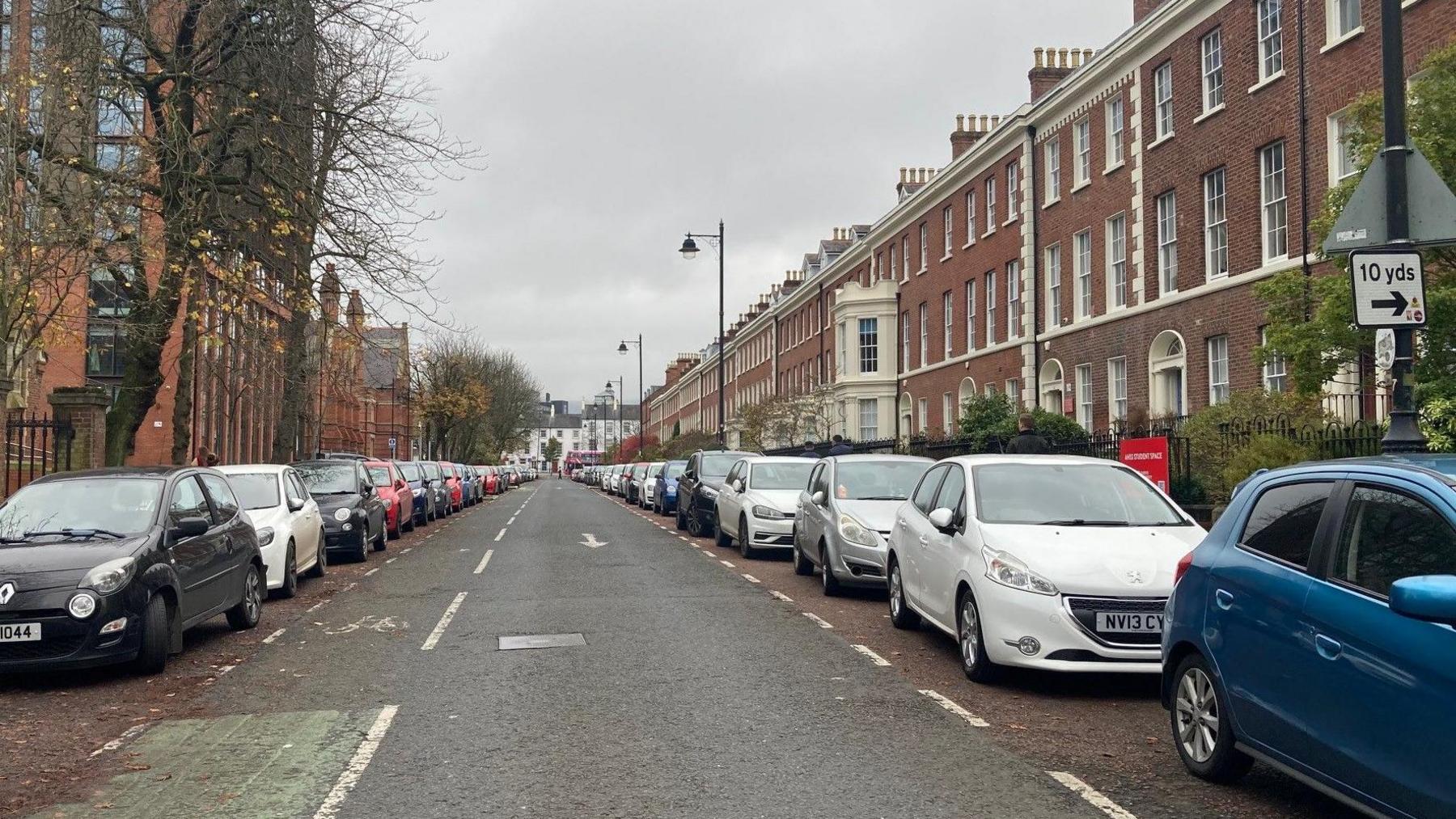 A road with cars parked on either side. The road is in-between a row of red-brick buildings, and a number of larger red-brick buildings. There are no visible spaces between the cars. 