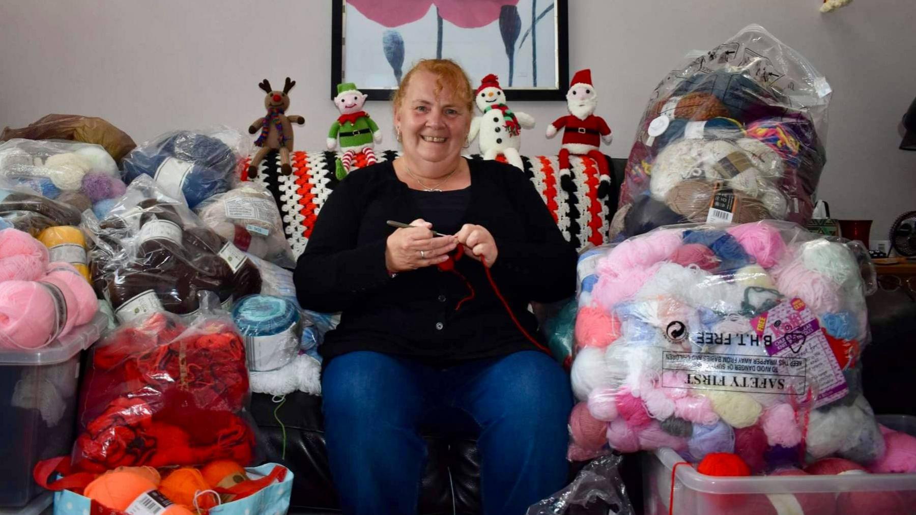 Rachel Williamson smiling on a sofa, holding knitting needles, surrounded by balls of wool