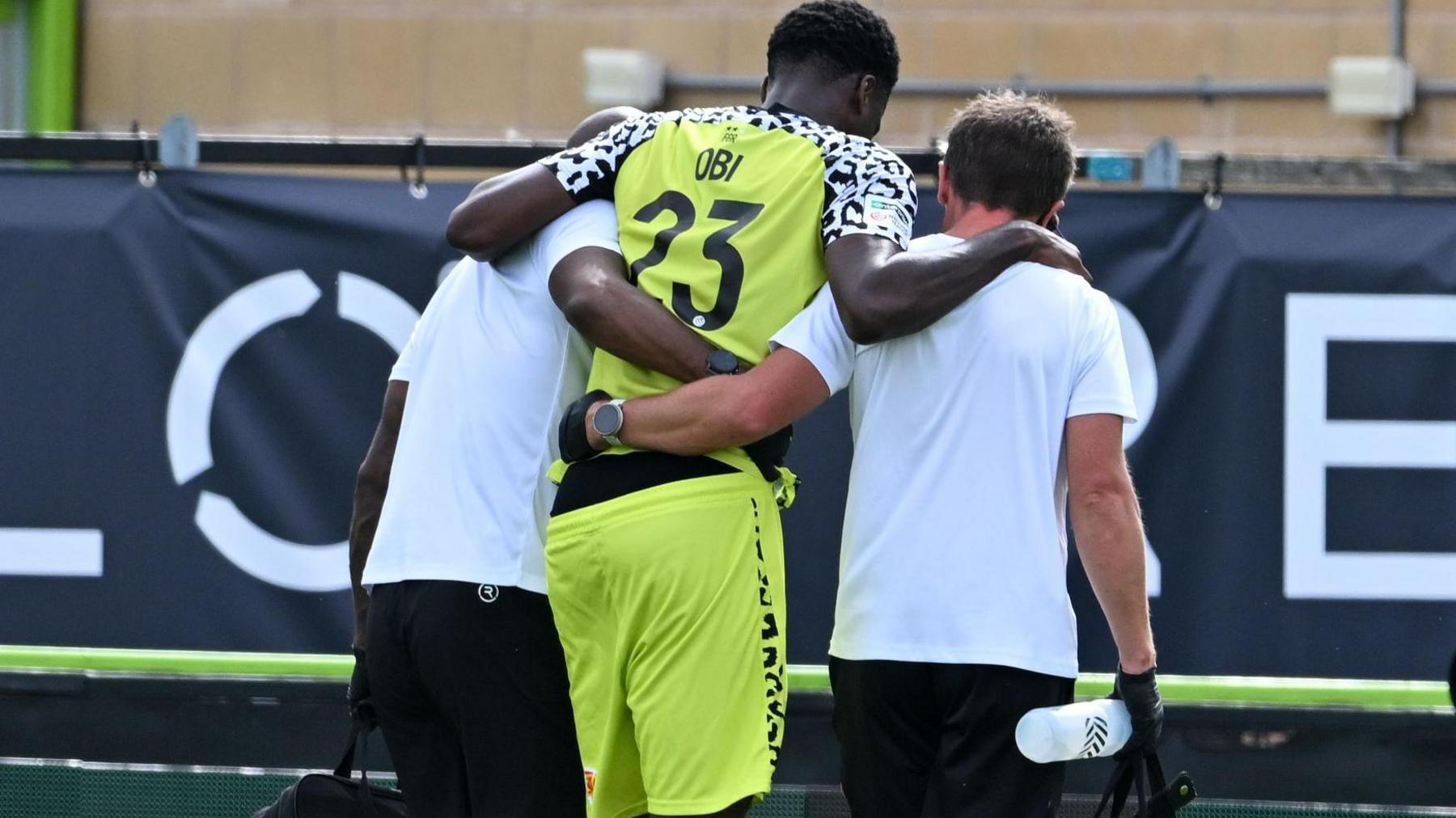 Emeka Obi in a pale yellow shirt and shorts being helped off by two men in white shirts who have their arms round him