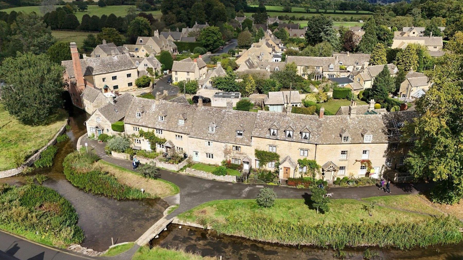 A quaint small village in the Cotswolds. In the foreground, there is a small river running perpendicular to a row of white stone houses with grey slate roofs and small windows. A tiny bridge stretches across the river, and is bordered either side by grassy patches of land. In the background, the houses are dotted between green spaces and trees.