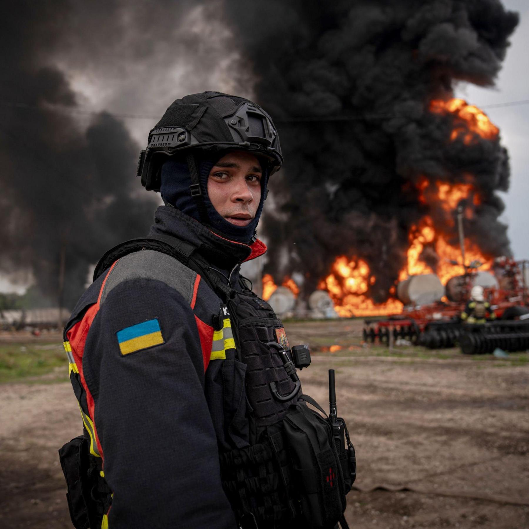 A member of the emergency services in north-eastern Sumy, dressed in high vis with the Ukraine flag sewn onto his arm and a combat helmet and balaclava, looks wearily at the camera while standing in front of a burning gas depot near the Russian border.