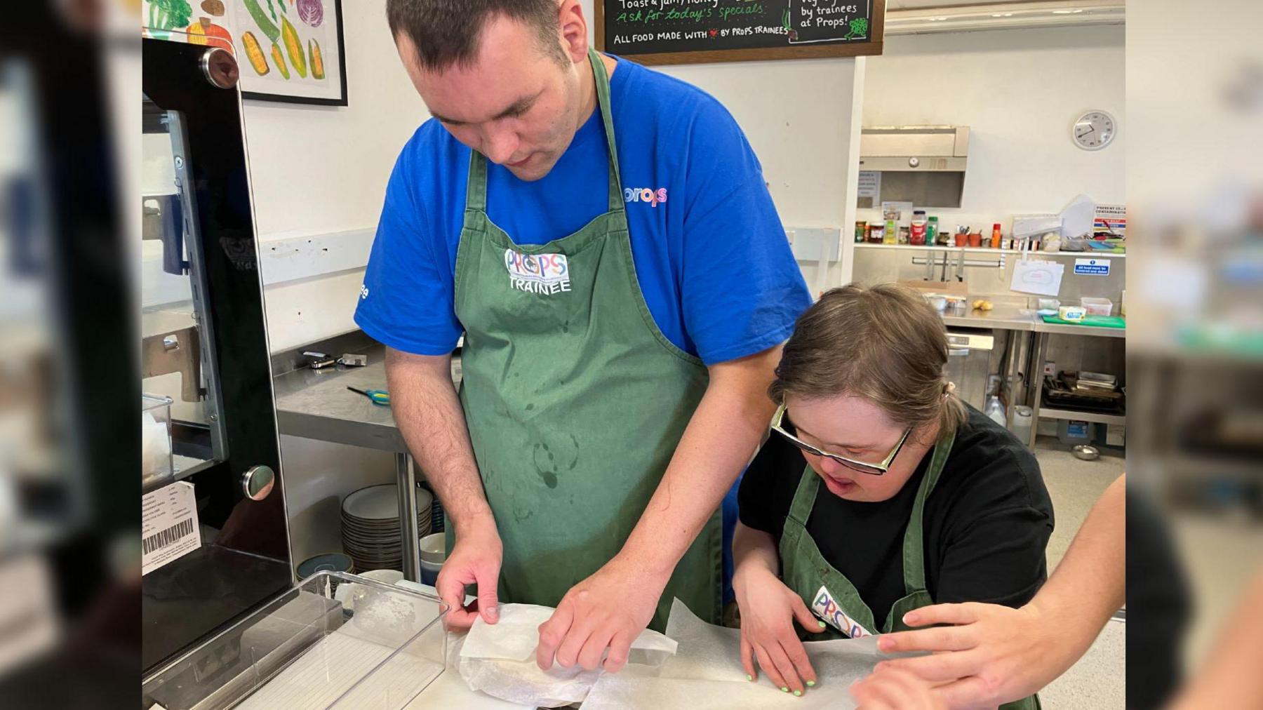 Two trainee chefs from Props wrapping sandwiches in greaseproof paper. They are both wearing green aprons and standing in a commercial kitchen. 