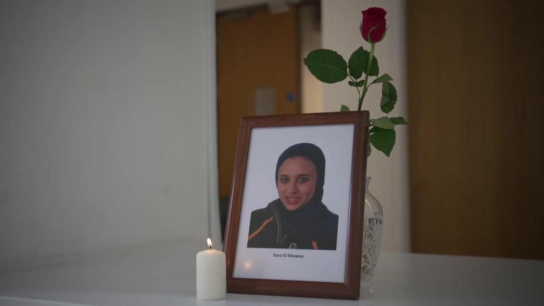 A framed headshot photo of Sara El-Khawas wearing a hijab. In front is a burning white candle, behind is a glass vase containing a single red rose.