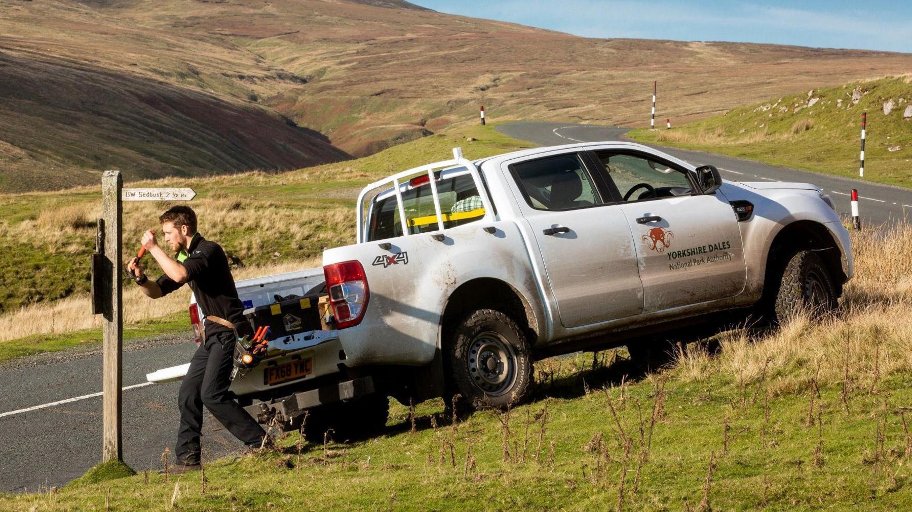 A man tries to fix a wooden sign on a road between moors with a white, mud-splattered 4x4 parked up on the side of the road