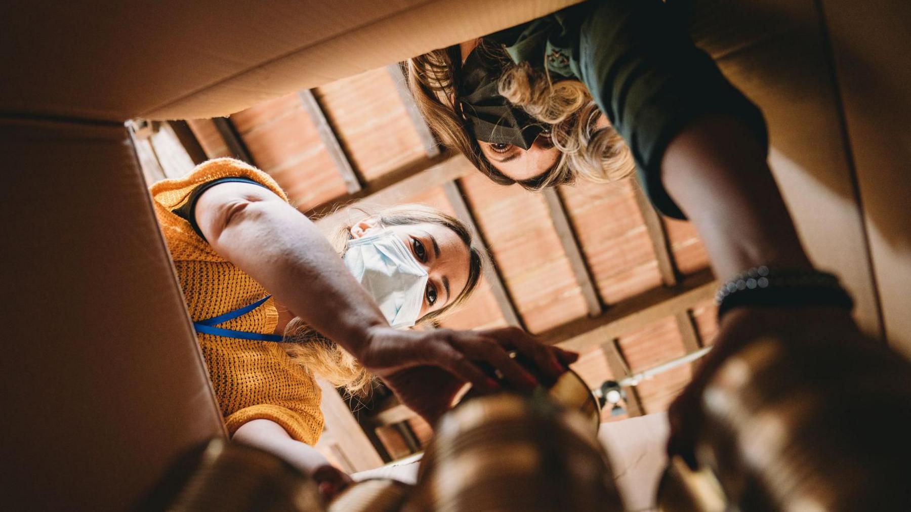 Volunteers are preparing a donation box with food - view from inside the cardboard box. People are wearing protective face masks.